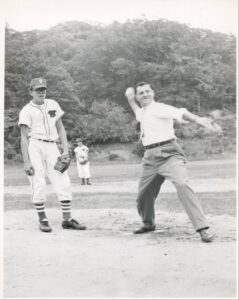 Adult pitching a ball.  Youth pitcher watching.  In the 1950’s.